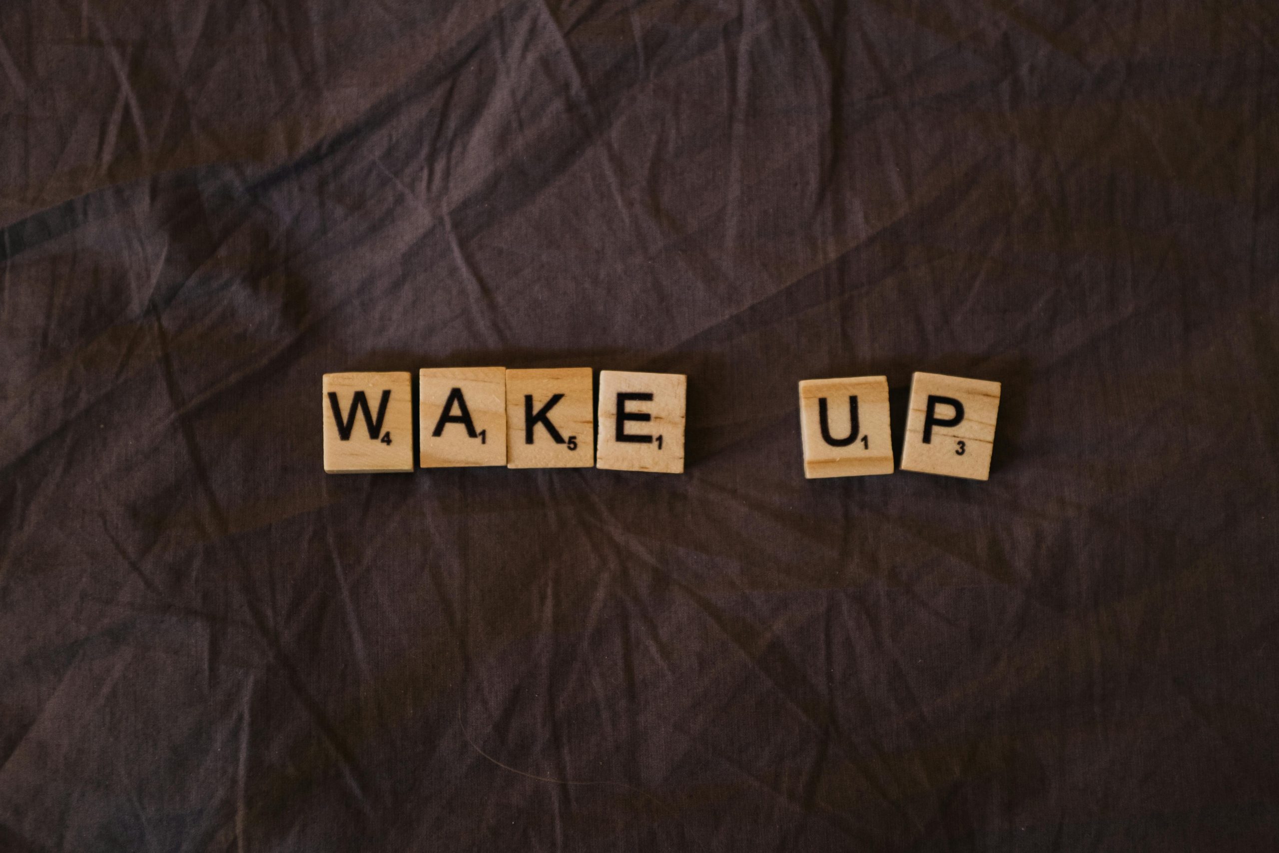 Wooden Scrabble tiles on a brown fabric surface spell out 'Wake Up', perfect for motivational themes.
